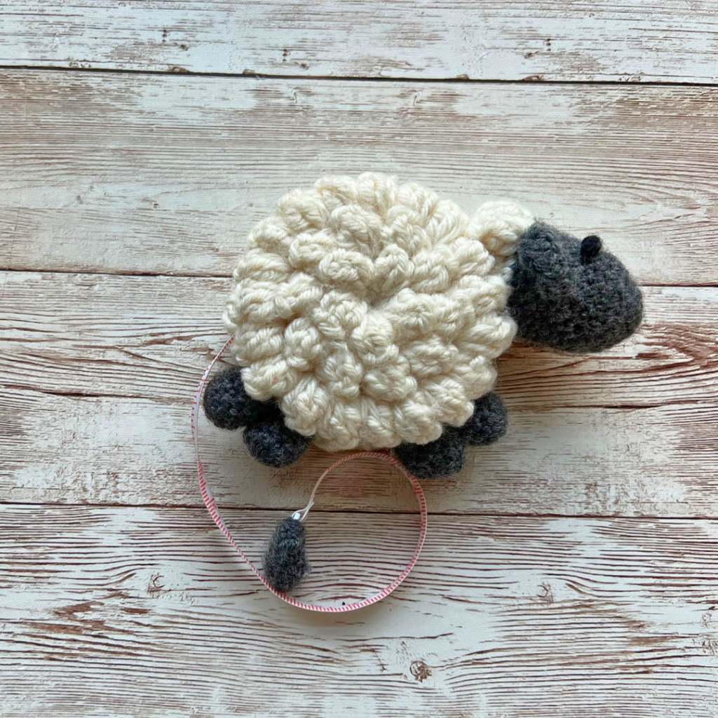 White Crochet Sheep tape measure laying on a wooden table facing to the right. 