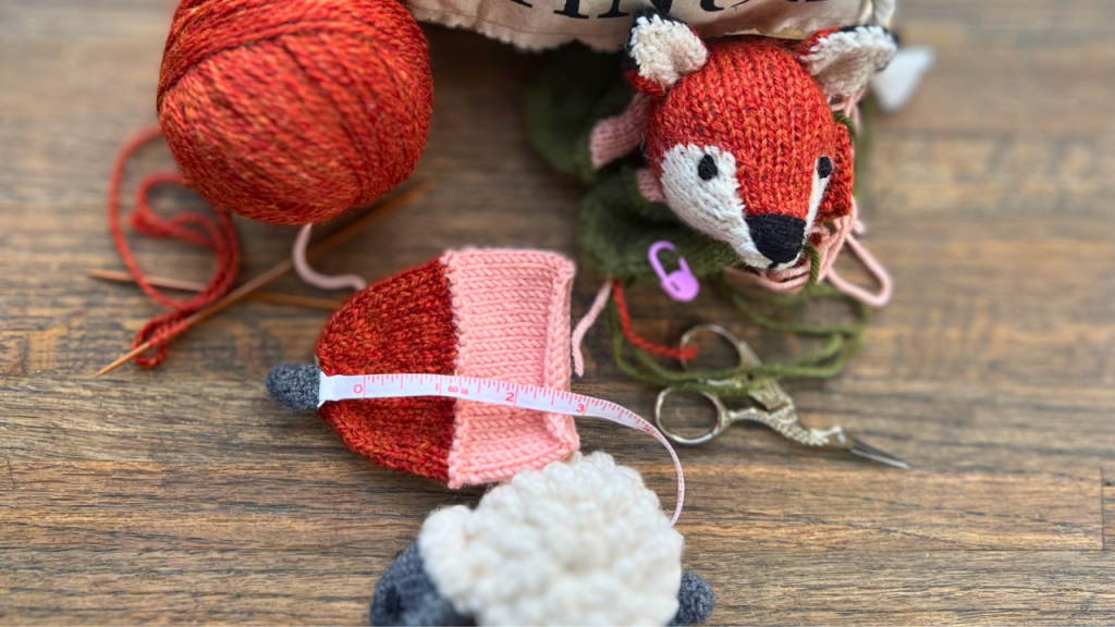 Ball of orange yarn with knitted toy fox head white and gray crochet sheep tape measure pair of scissors and a notion