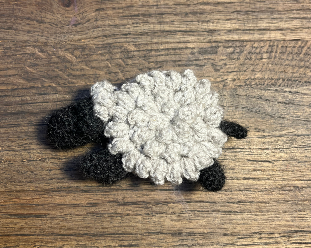 White crochet sheep tape measure laying on a wooden table facing to the left with the tail in 